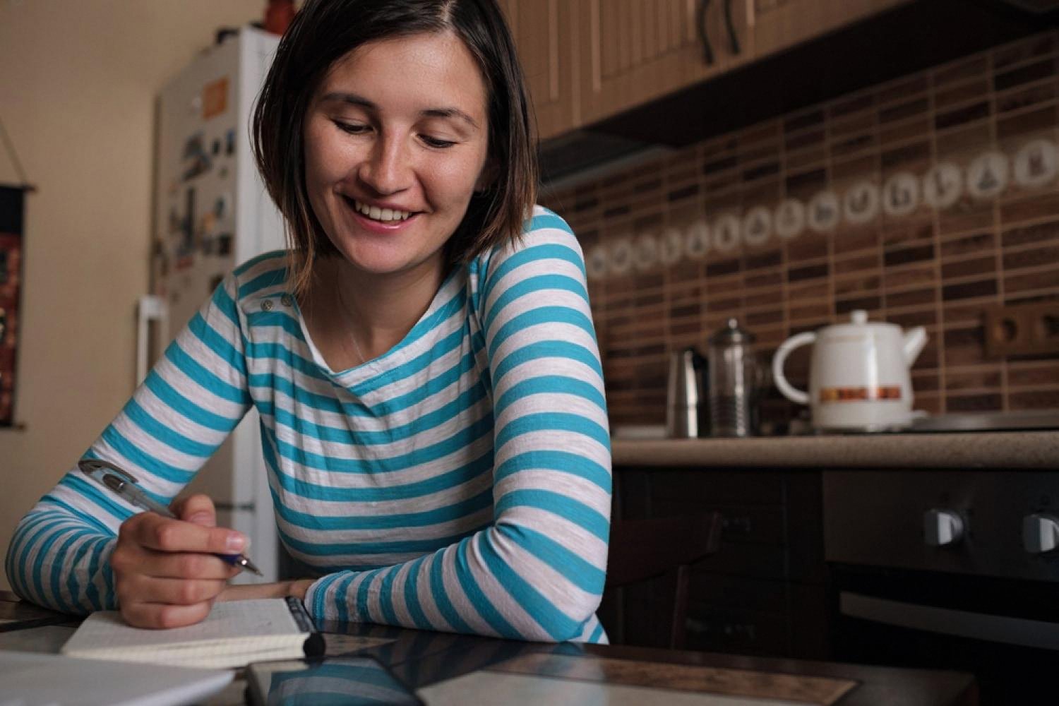 Young woman smiling while writing in a notebook at her kitchen table, planning a personal budget